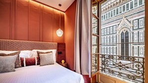 Habitación de hotel con una cama grande, un cabecero decorativo, una lámpara colgante y vistas a un edificio de arquitectura intrincada a través de una ventana con barandilla de metal.