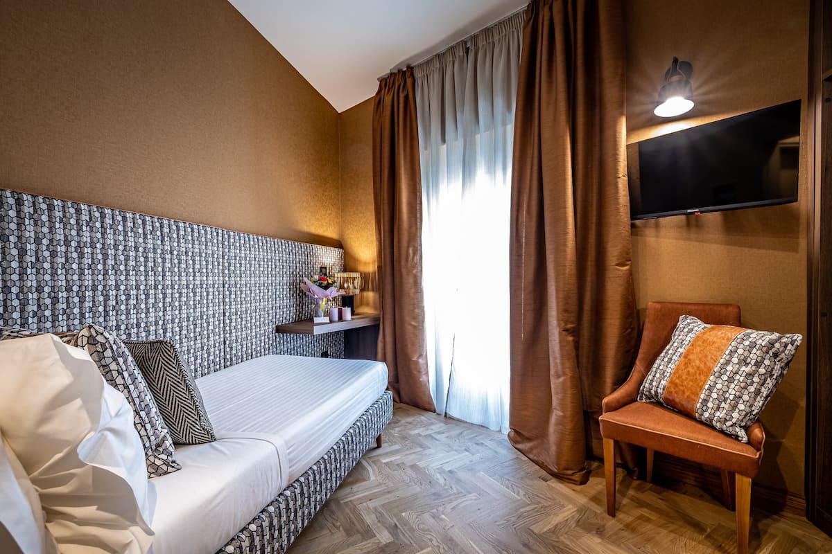 Habitación de hotel con una cama grande, una silla, un televisor montado en la pared y un ventanal con cortinas.