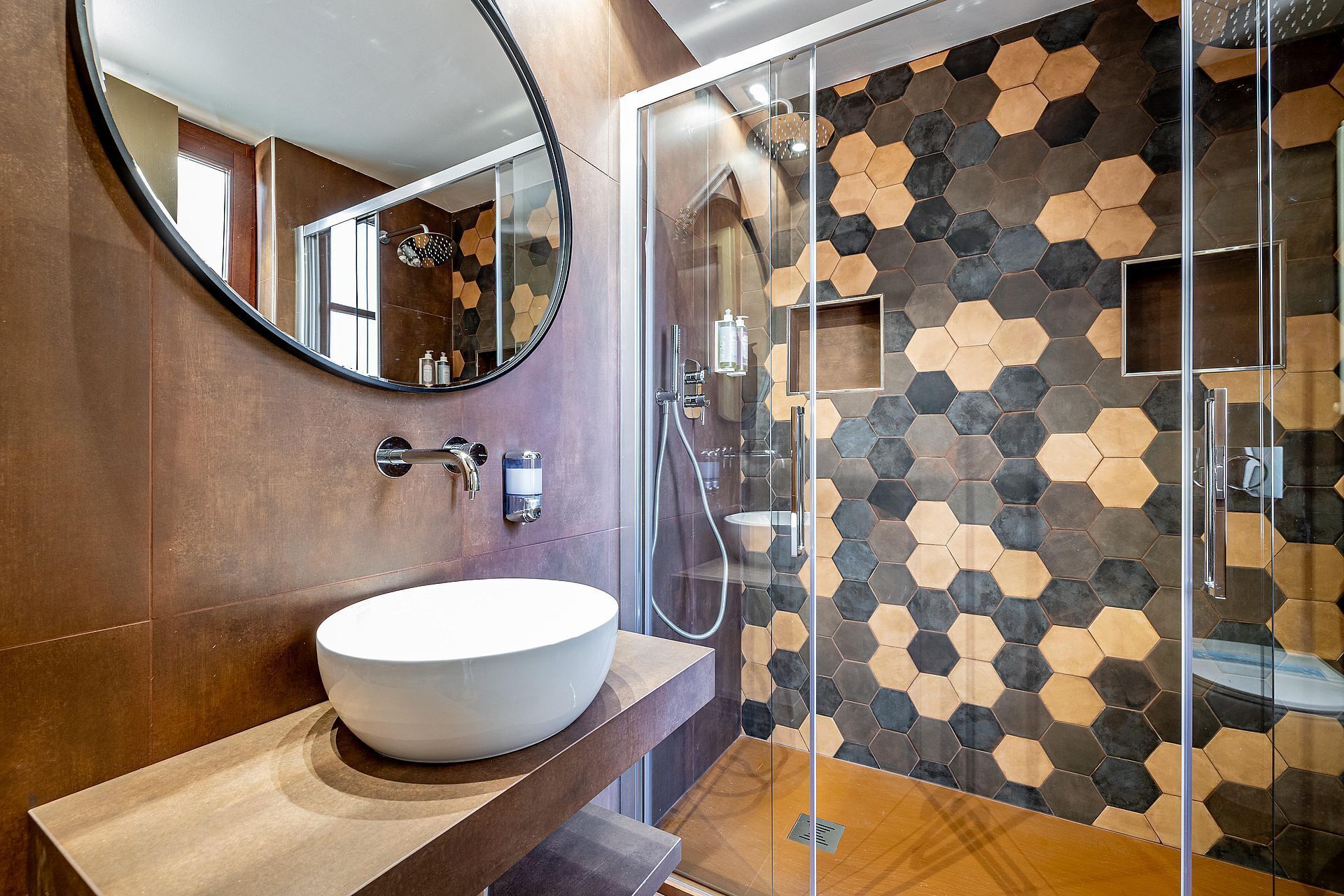 A modern bathroom with a round mirror, a white vessel sink, and a shower area with a hexagonal tile wall.