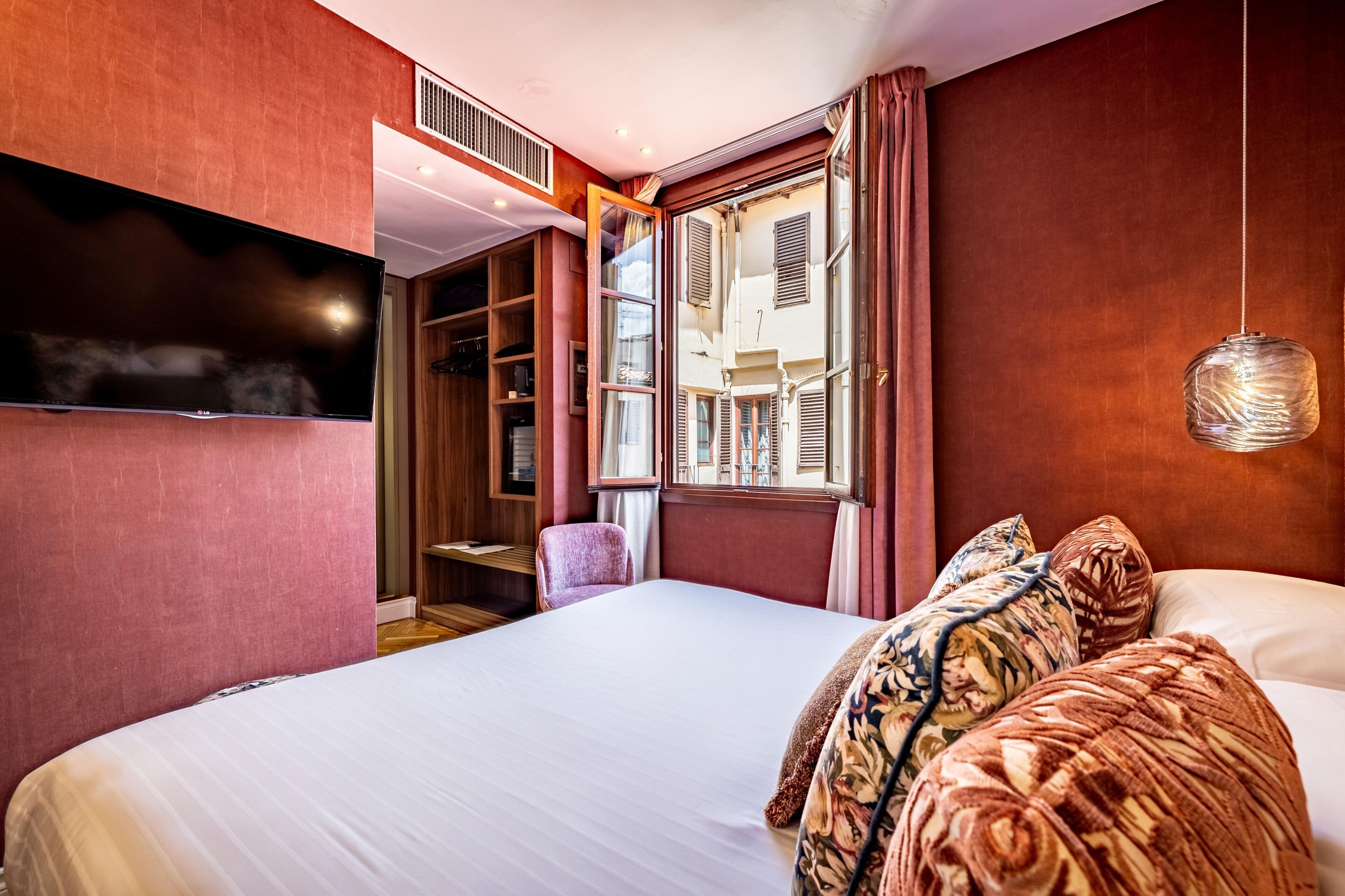 A hotel room with a large bed, a television mounted on the wall, a window with shutters, and a hanging light fixture.