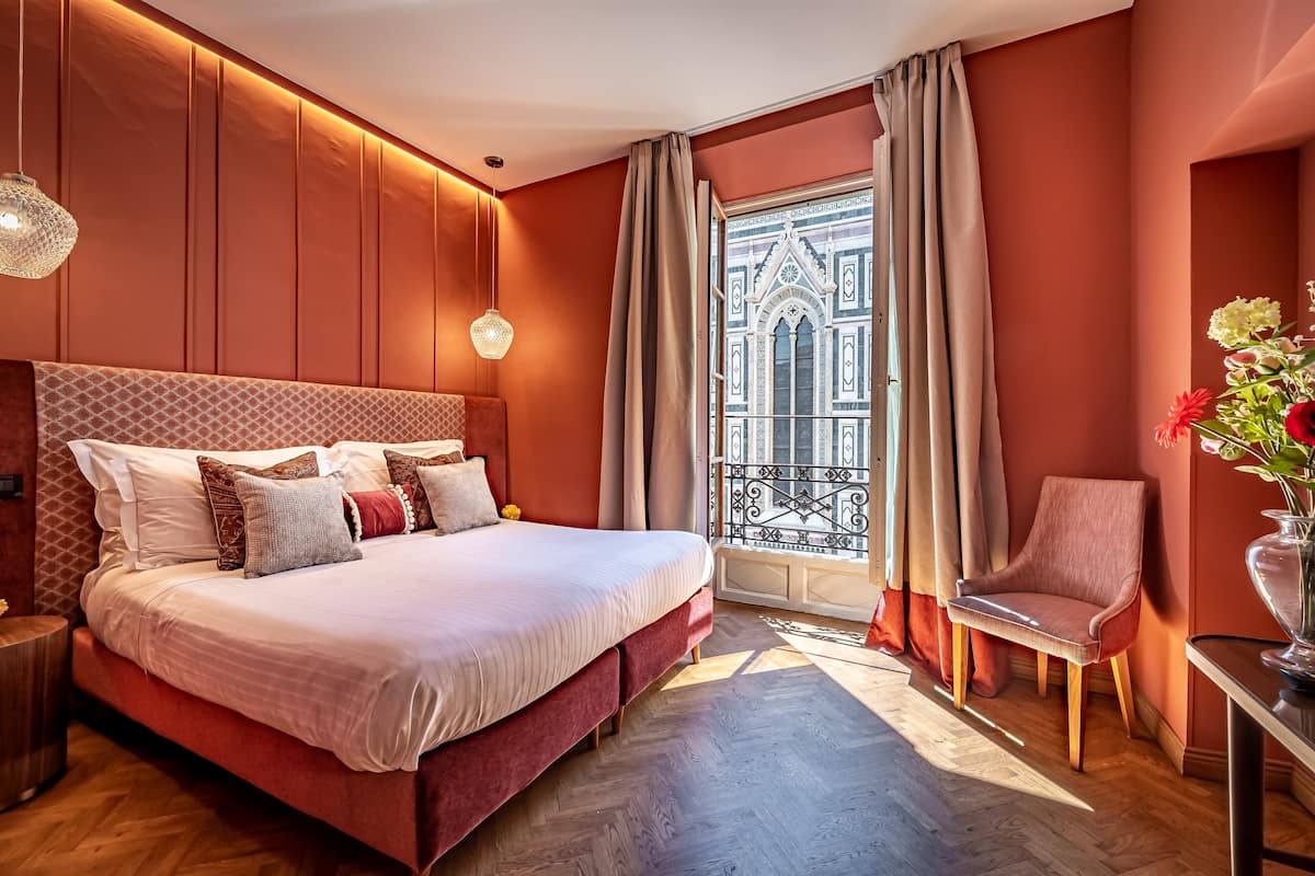 Un dormitorio con una cama grande, una silla, una mesa con un florero y una ventana con cortinas.
