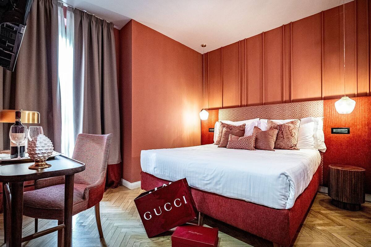 Una habitación de hotel con una cama grande, un escritorio con una lámpara y un bolso de Gucci rojo en el suelo.