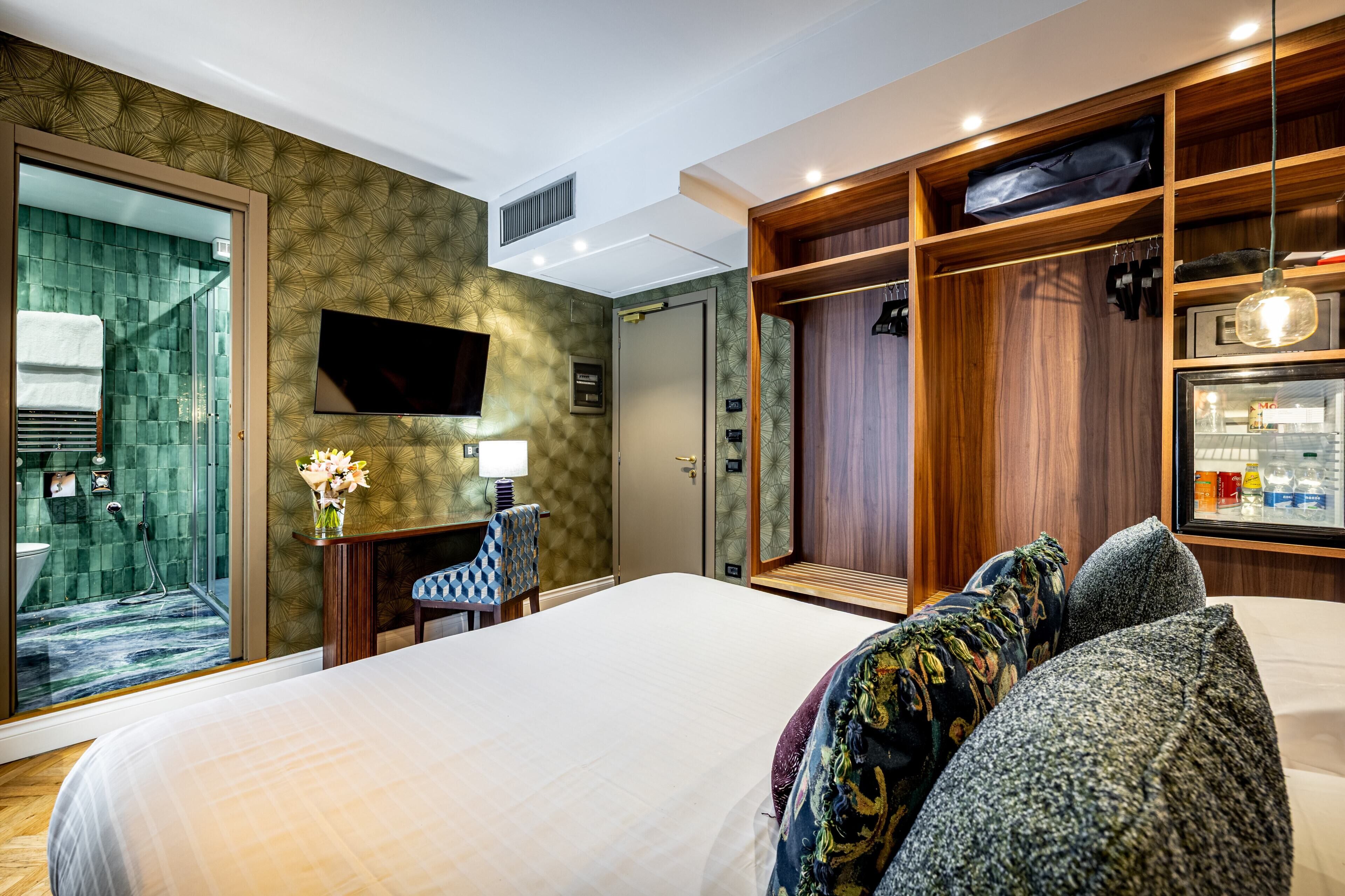 A hotel room with a bed, a desk with a lamp, a TV, a walk-in closet, and a shower area.