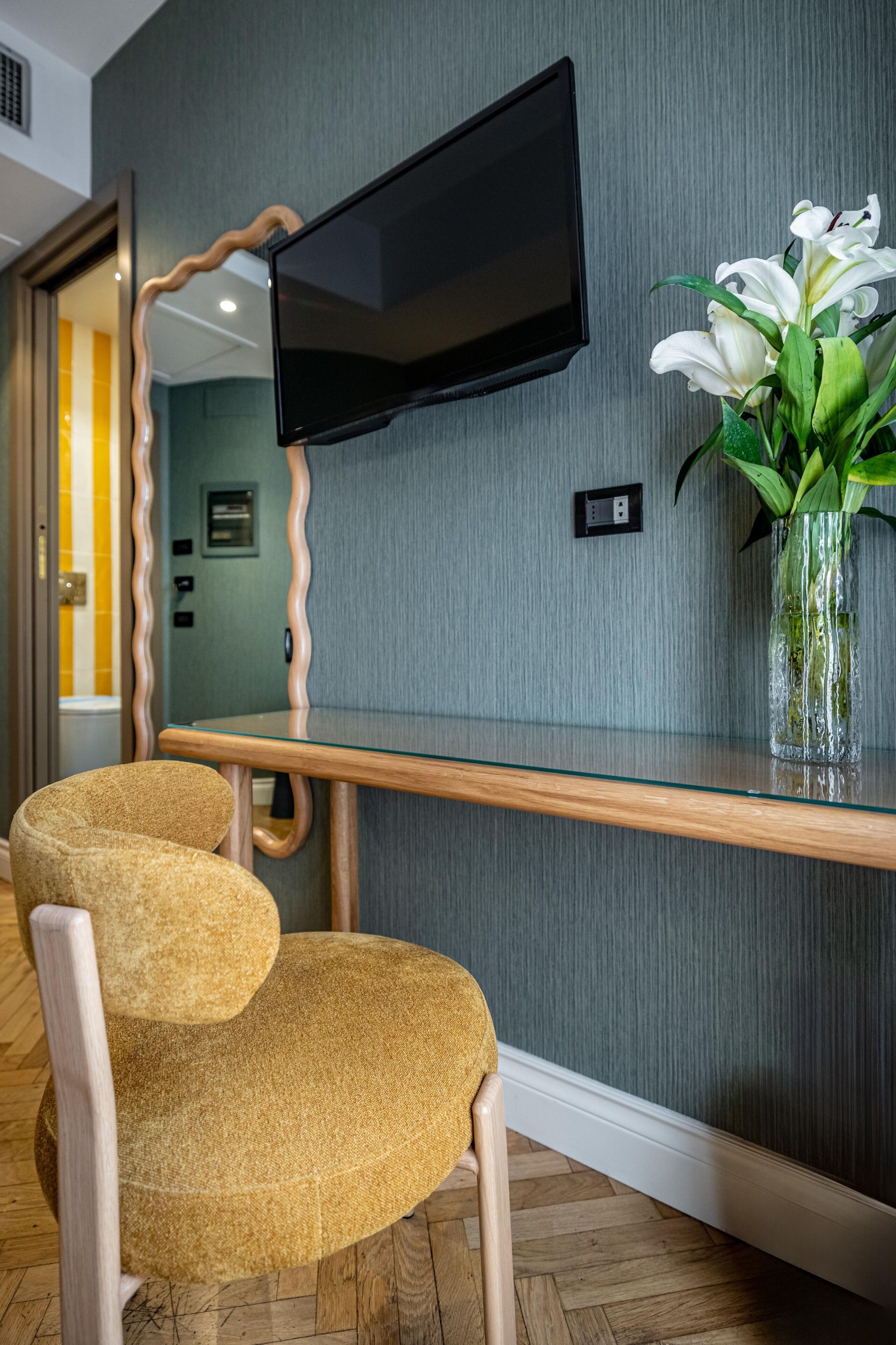 A modern room with a flat-screen TV mounted on a textured wall, a wooden desk with a vase of white flowers, and a pair of upholstered chairs.