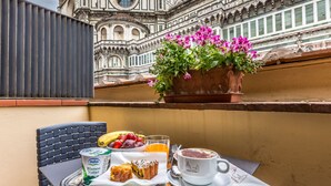 Una mesa con alimentos para el desayuno, una planta en maceta con flores y vistas a un edificio histórico.