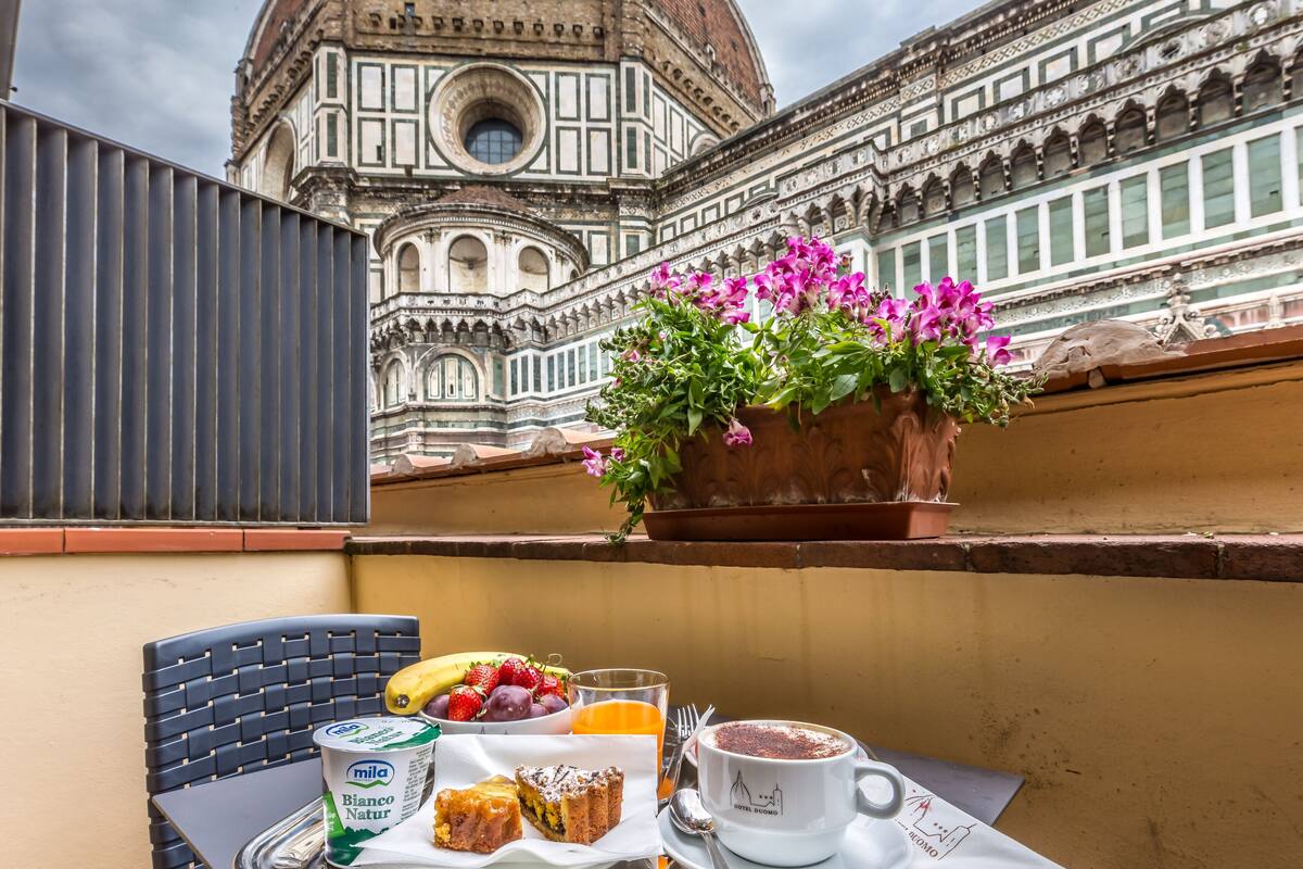 Una mesa con alimentos para el desayuno, una planta en maceta con flores y vistas a un edificio histórico.