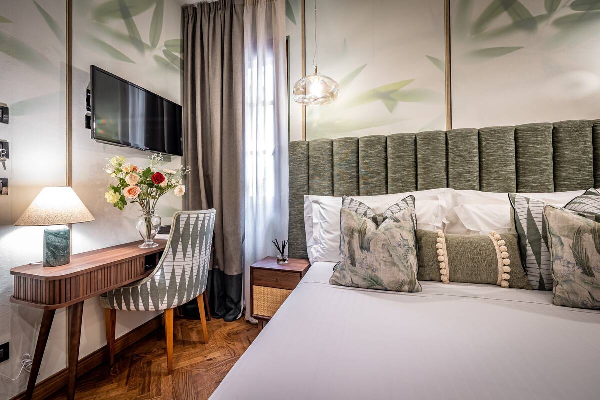 Habitación de hotel con cama, escritorio, silla, televisor y pared con estampado de hojas.