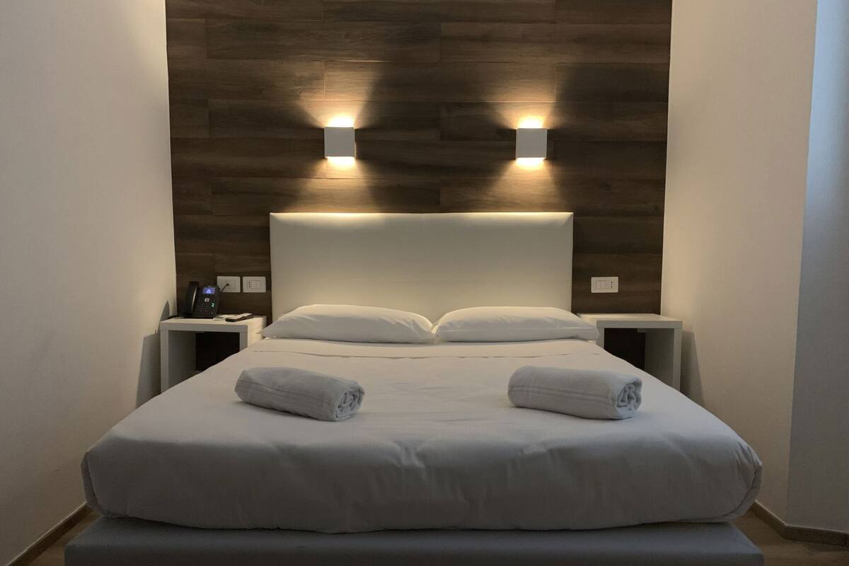 A hotel room with a large bed, two bedside tables, a wooden headboard, and two wall-mounted lights.