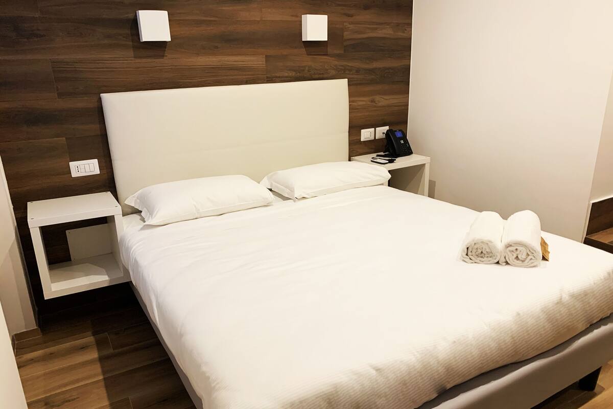 A neatly made bed with white linens and pillows, a bedside table with a phone, and a wall-mounted light fixture.