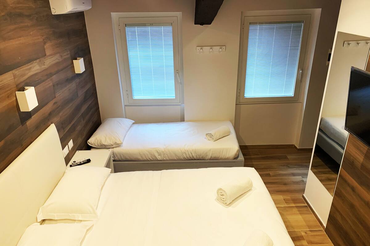 A hotel room with two beds, wooden flooring, a TV, and windows with blinds.