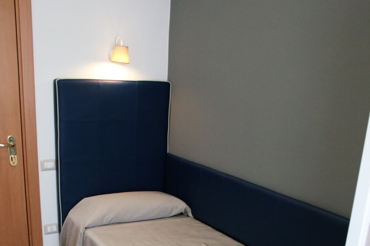 Quarto pequeno com uma cama de solteiro, porta de madeira, mesa de cabeceira e luminária na parede.