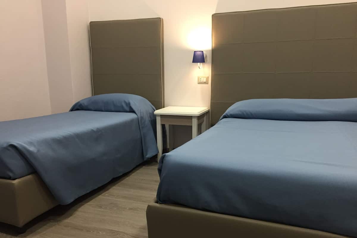 Quarto de hotel com duas camas de solteiro, uma mesinha e uma luminária presa à parede.