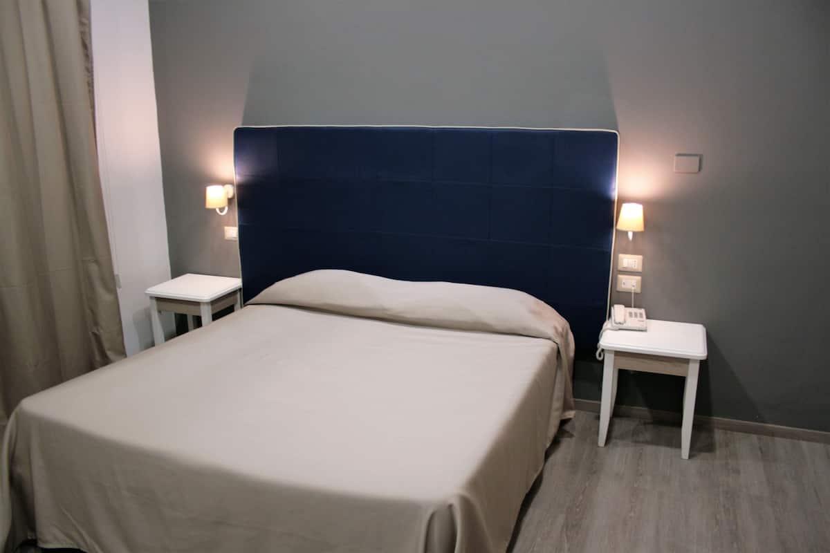 Cama bem arrumada com cabeceira escura, duas mesas de cabeceira com abajures e cortina em tom claro.