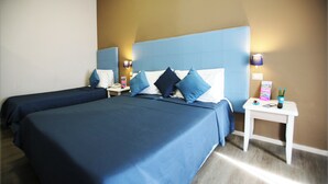 A hotel room with two beds, each with blue bedding and pillows, a nightstand with a lamp, and a small table with a remote control and a book. - Hotel Magenta (Florence)