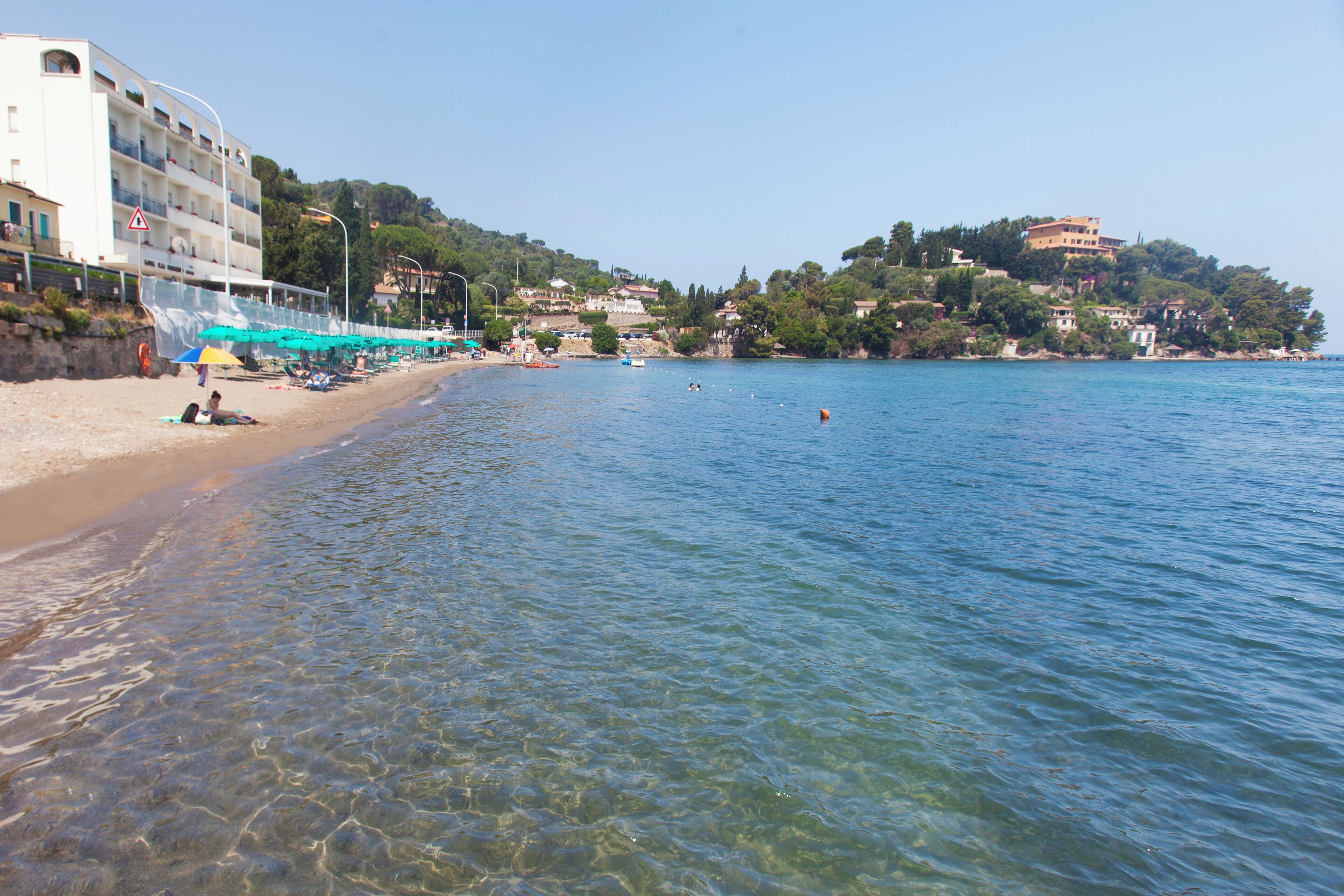 A beach with clear water, umbrellas, and a building with multiple floors.