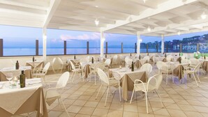 An outdoor dining area with tables and chairs, large windows with a view of the sea, and hanging lights. - Baia d'Argento (Monte Argentario)