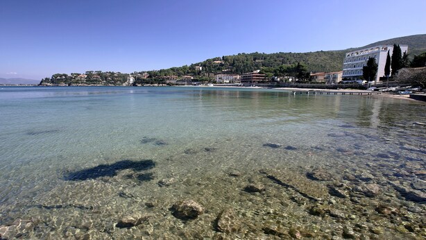 A coastal view with clear shallow waters, rocky seabed, and buildings on a hillside. - Baia d'Argento (Monte Argentario)