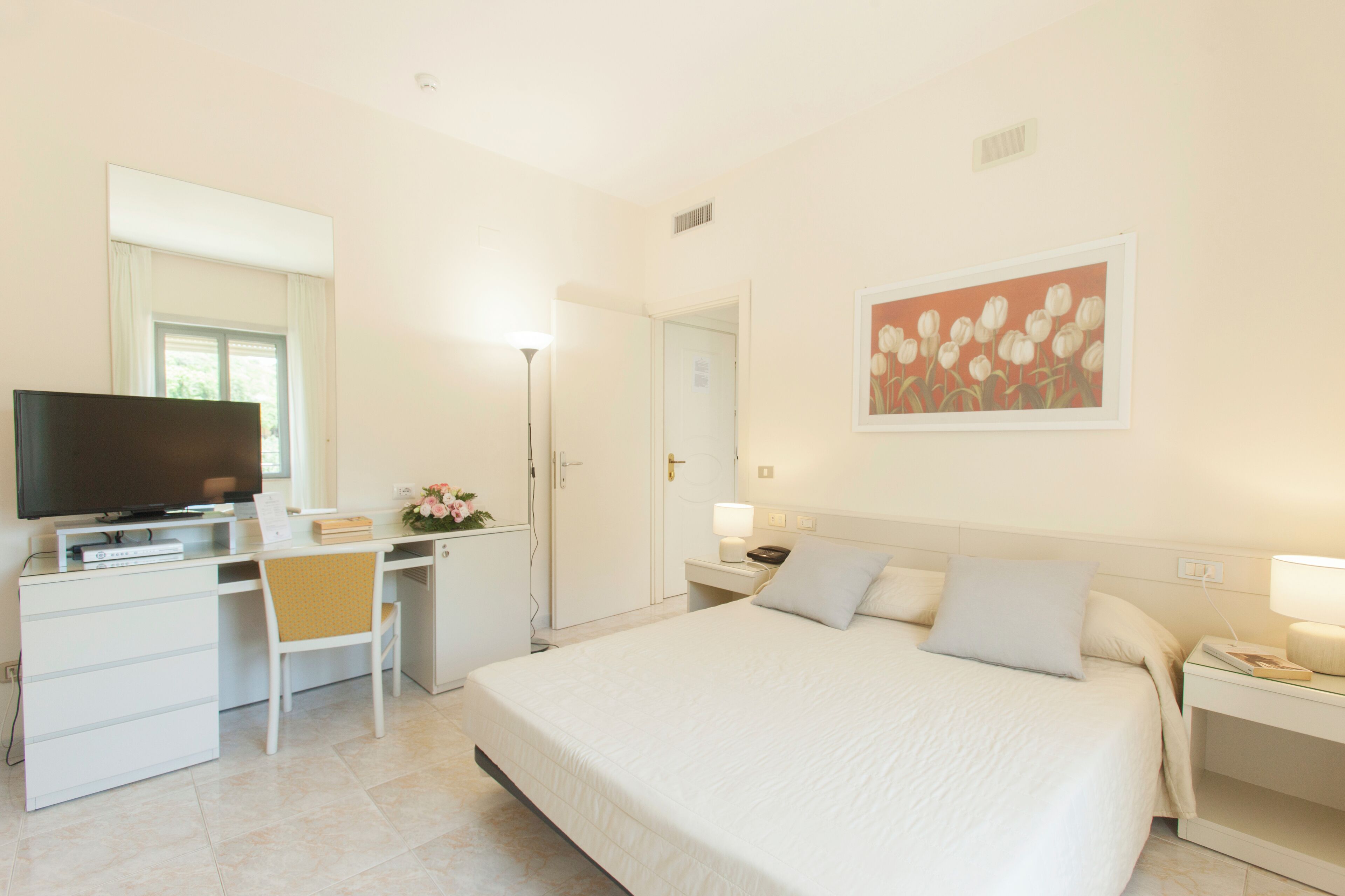 A hotel room with a bed, a desk with a TV, a chair, a nightstand, and a wall-mounted painting.