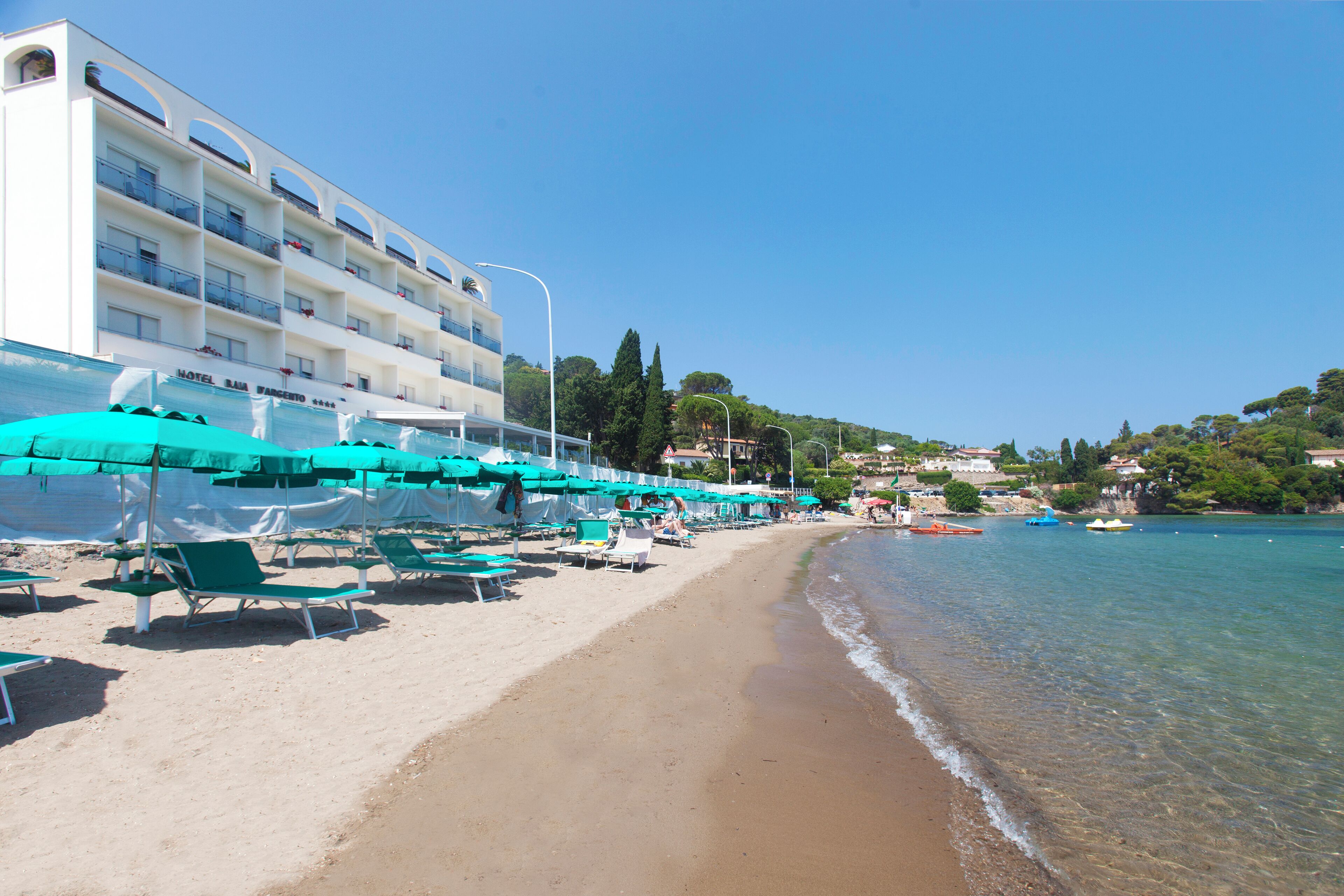 A beach with rows of green lounge chairs and umbrellas, a hotel building with balconies, and clear blue water.