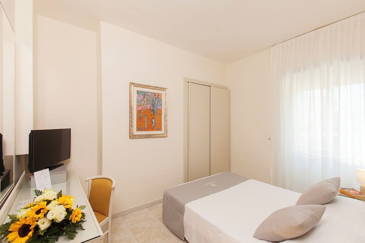 Une chambre d’hôtel avec un lit, un bureau avec un ordinateur, une chaise, un tableau encadré au mur et une fenêtre avec des rideaux.