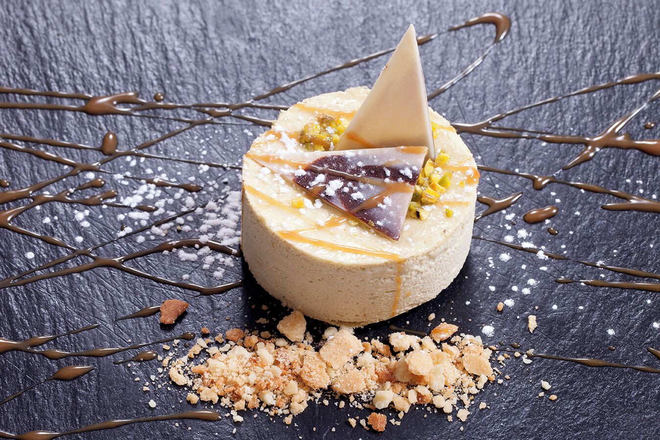 A dessert with a cone on top, drizzled with chocolate, and surrounded by crumbs and sugar on a dark surface.