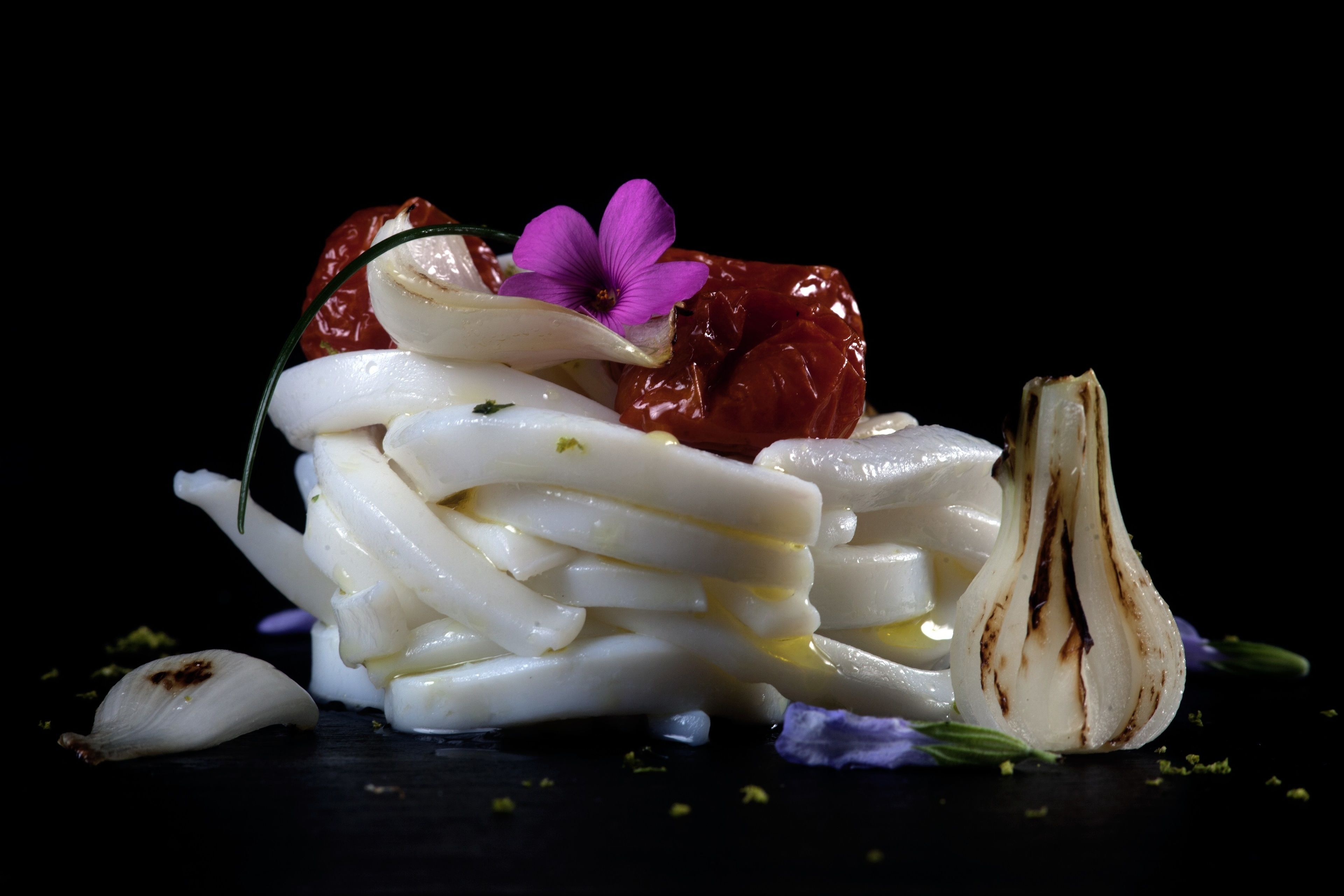 A dish with white noodles, a red chili, a purple flower, and a grilled vegetable.