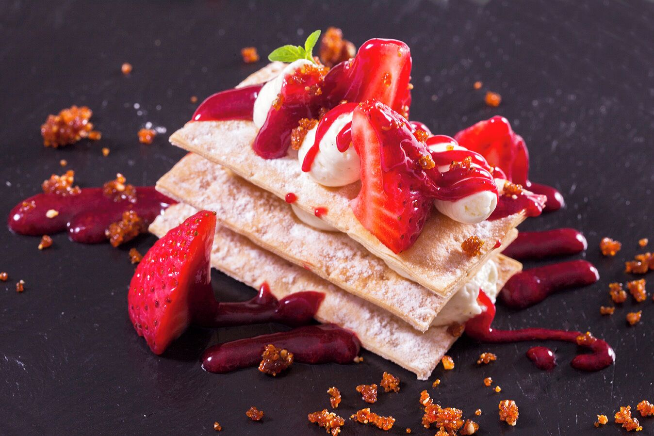 Una pila de panqueques delgados y crujientes cubiertos con fresas frescas, crema batida y salsa de frutos rojos, con migas esparcidas alrededor.