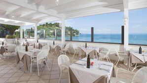 An outdoor dining area with white tables and chairs, overlooking a scenic coastal view. - Baia d'Argento (Monte Argentario)
