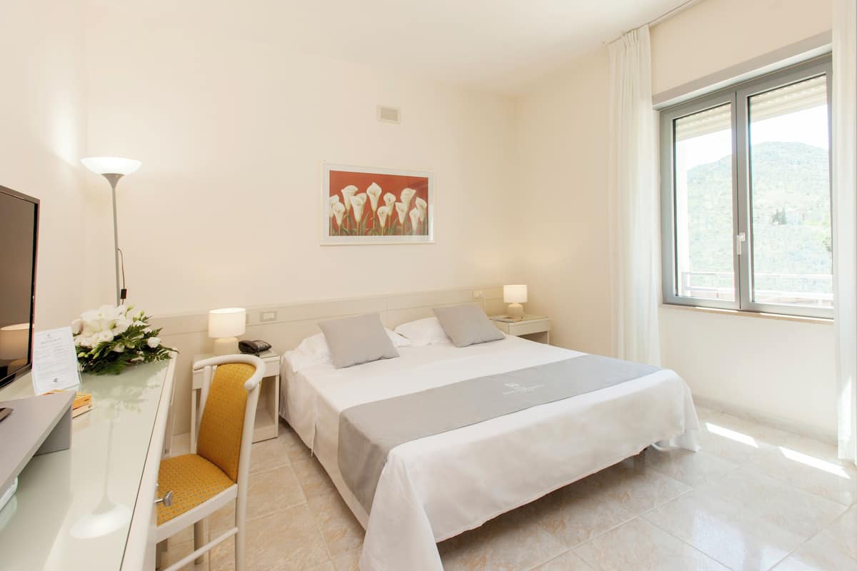 Une chambre d’hôtel comprenant un lit, un bureau avec un ordinateur, une chaise, une table de chevet, une lampe et une fenêtre donnant sur une vue sur la montagne.