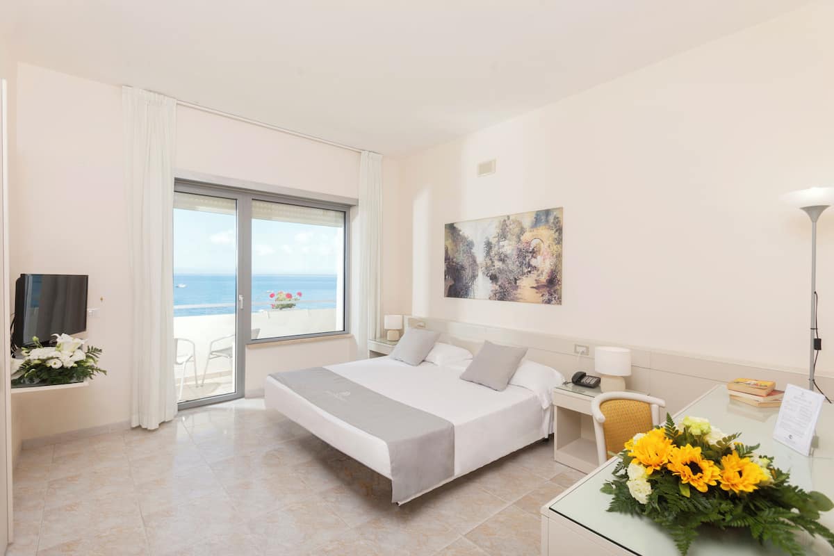 Une chambre d’hôtel comprenant un lit, un bureau avec un téléphone, une chaise, une lampe de bureau et une vue sur la mer, accessible par une porte coulissante.