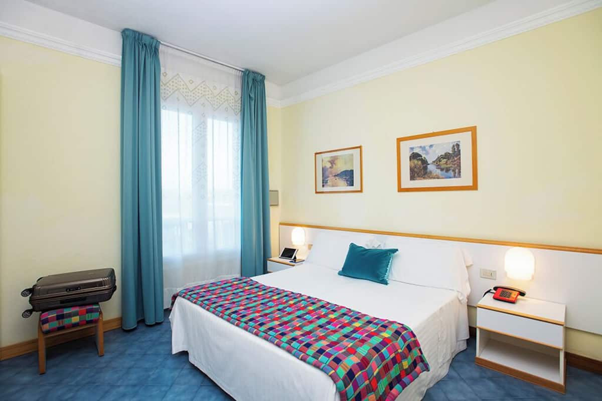 A hotel room with a bed, a nightstand with a lamp, a suitcase, and two framed pictures on the wall.