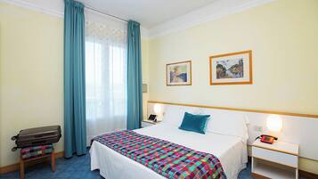 Camera d'albergo con un letto, un comodino con una lampada, una valigia e due quadri appesi al muro.