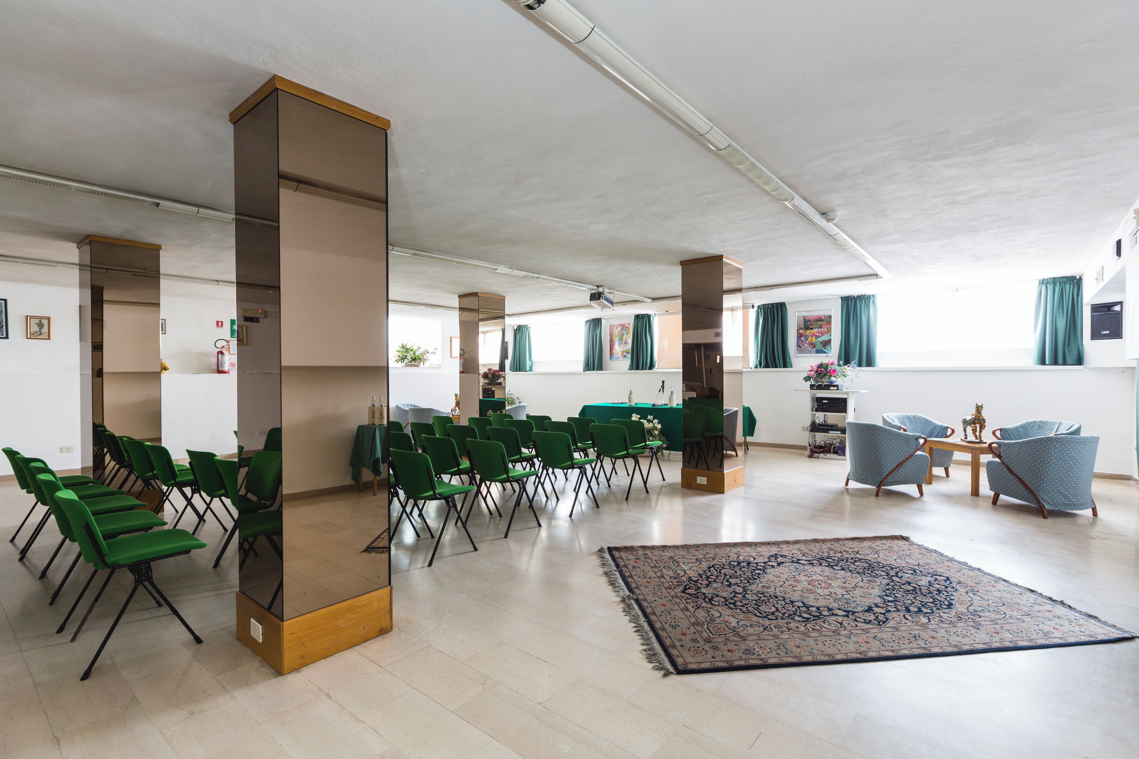 A spacious room with green chairs arranged in rows, a large patterned rug, and a small table with chairs.