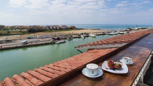 Vista al mar desde una terraza con una mesa lista para comer, con vistas a un puerto deportivo con barcos y una playa en el fondo.