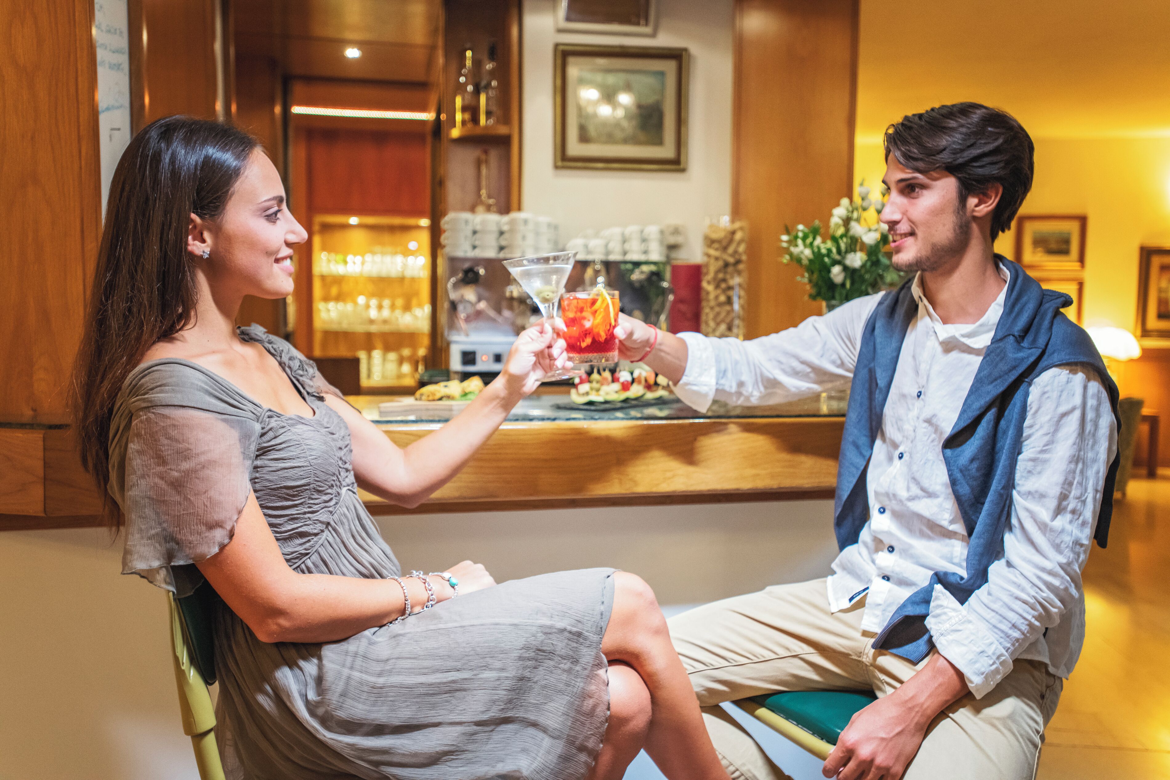 Un couple est assis au bar du hall d’un hôtel. On y voit un comptoir, des étagères remplies de verres et un tableau encadré accroché au mur.