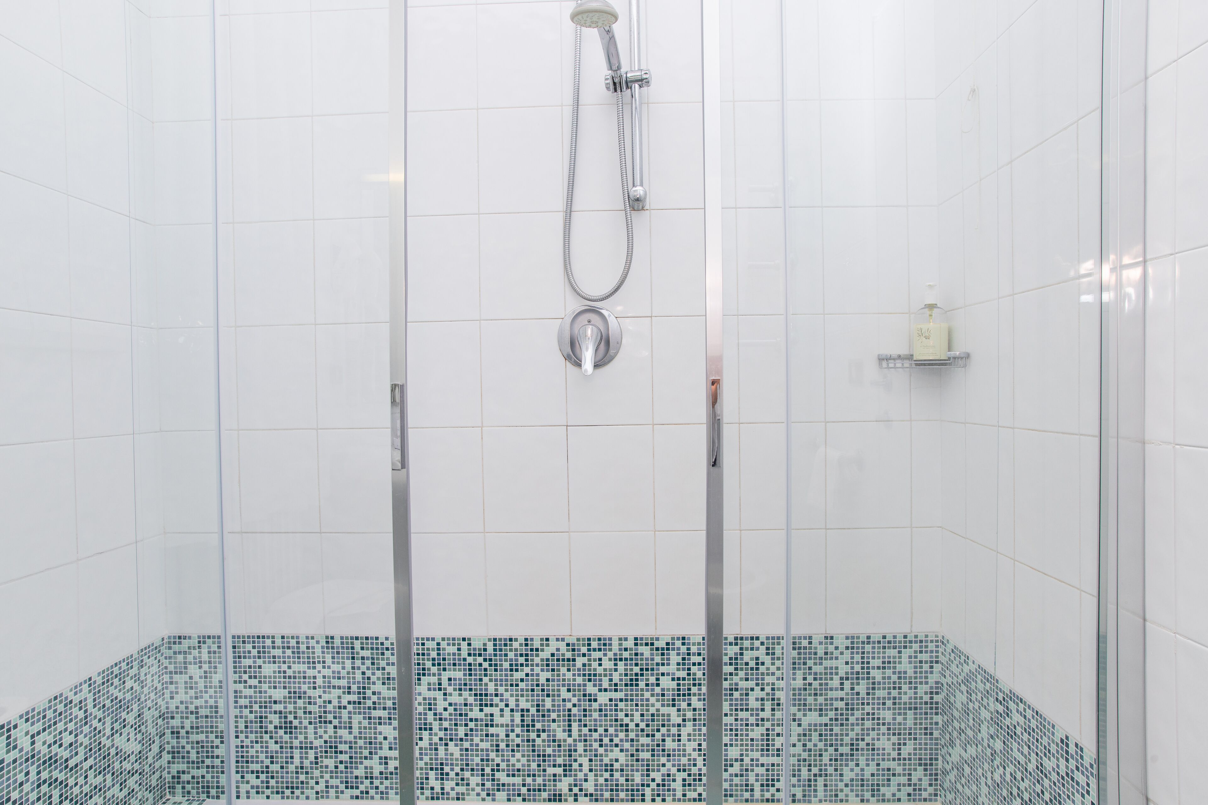 Une douche avec une porte en verre, un mur carrelé et une petite étagère sur laquelle se trouve une bouteille.