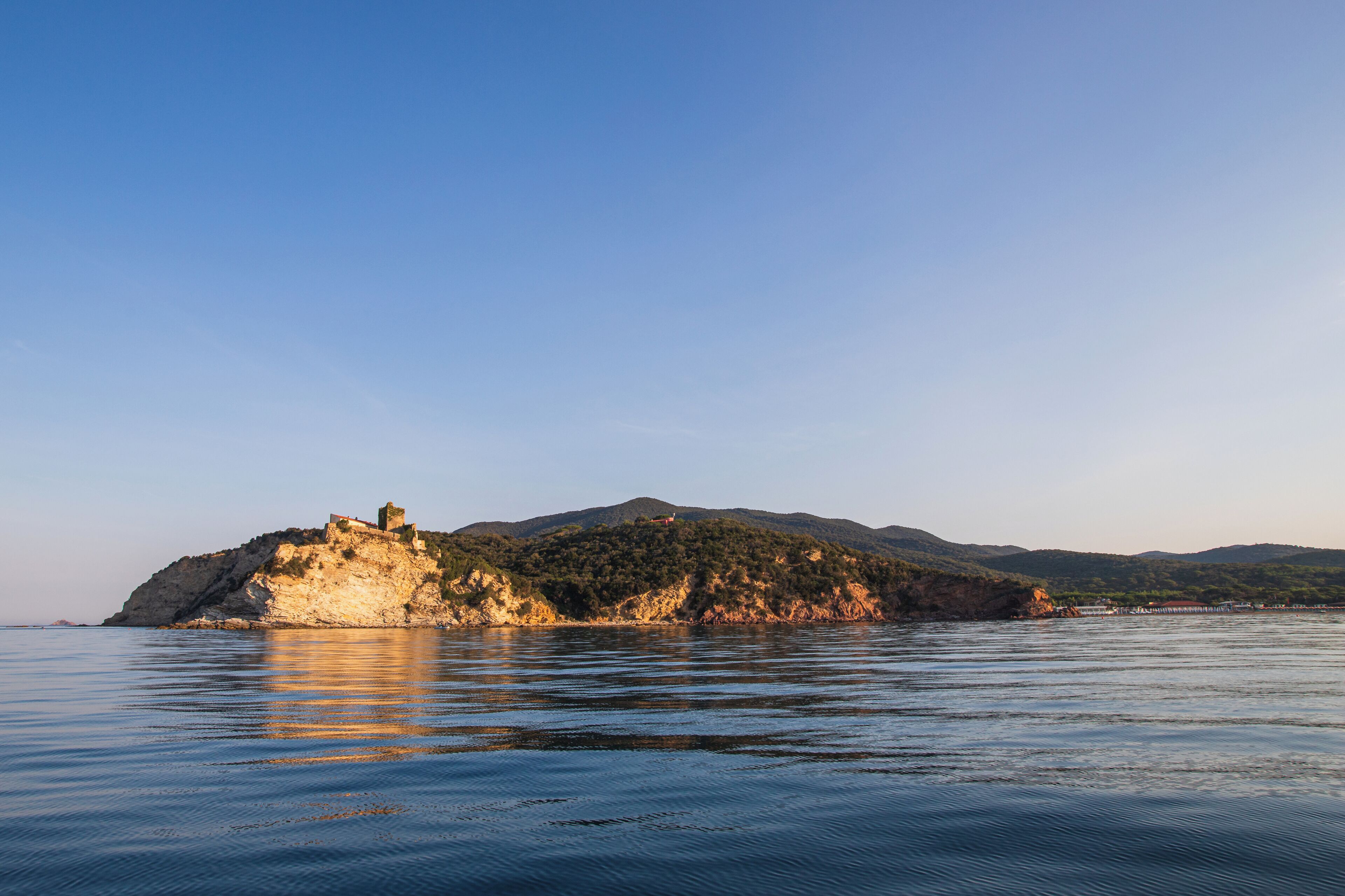 A coastal landscape with a rocky island featuring a small building, calm water, and distant hills.