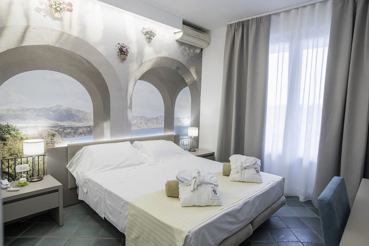 A hotel room with a large bed, two bedside tables, a nightstand, and a view of mountains through arched windows.