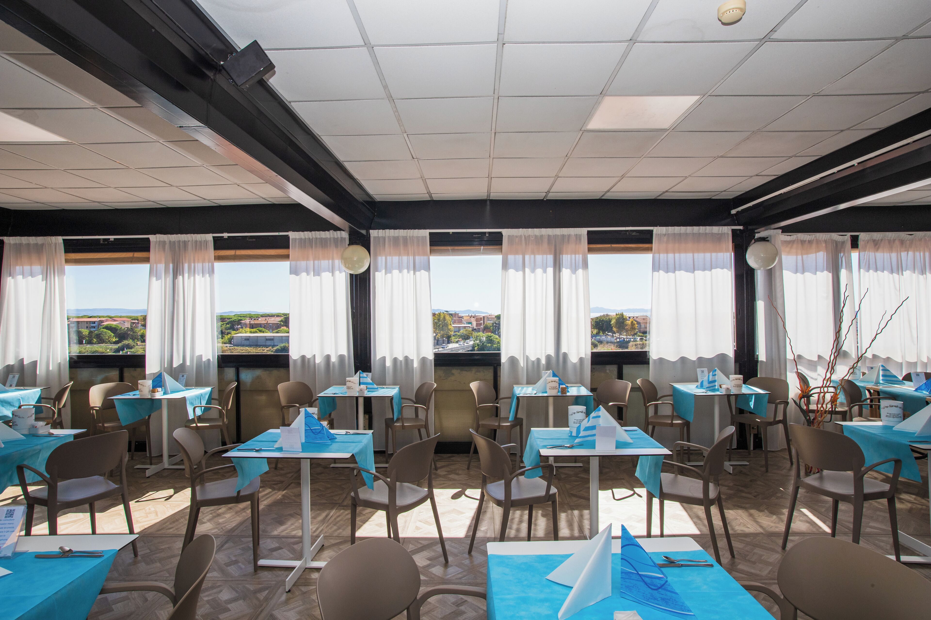 A restaurant with multiple tables and chairs, large windows with curtains, and a view of the cityscape.
