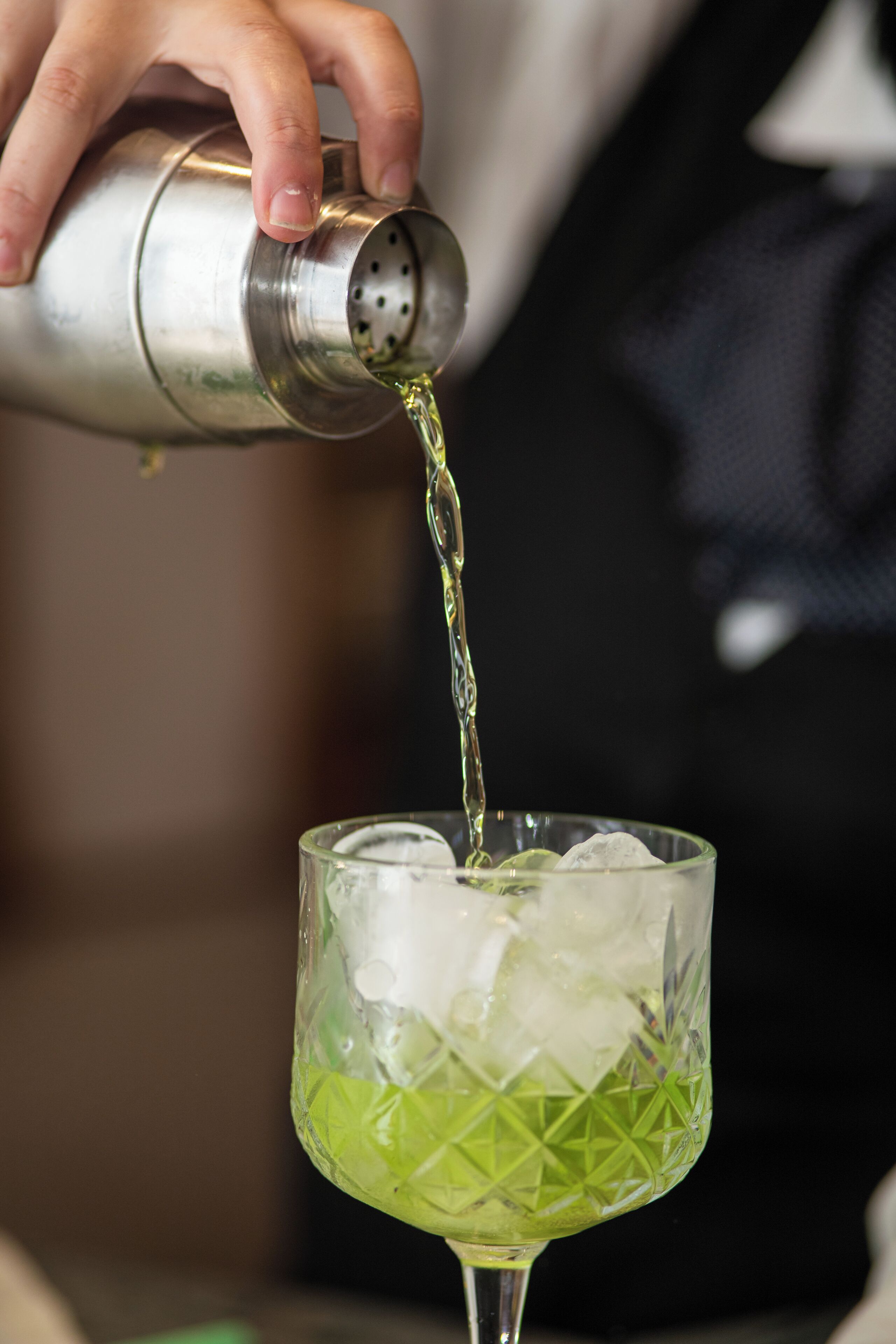 A hand pouring a clear liquid from a metal shaker into a glass with ice.