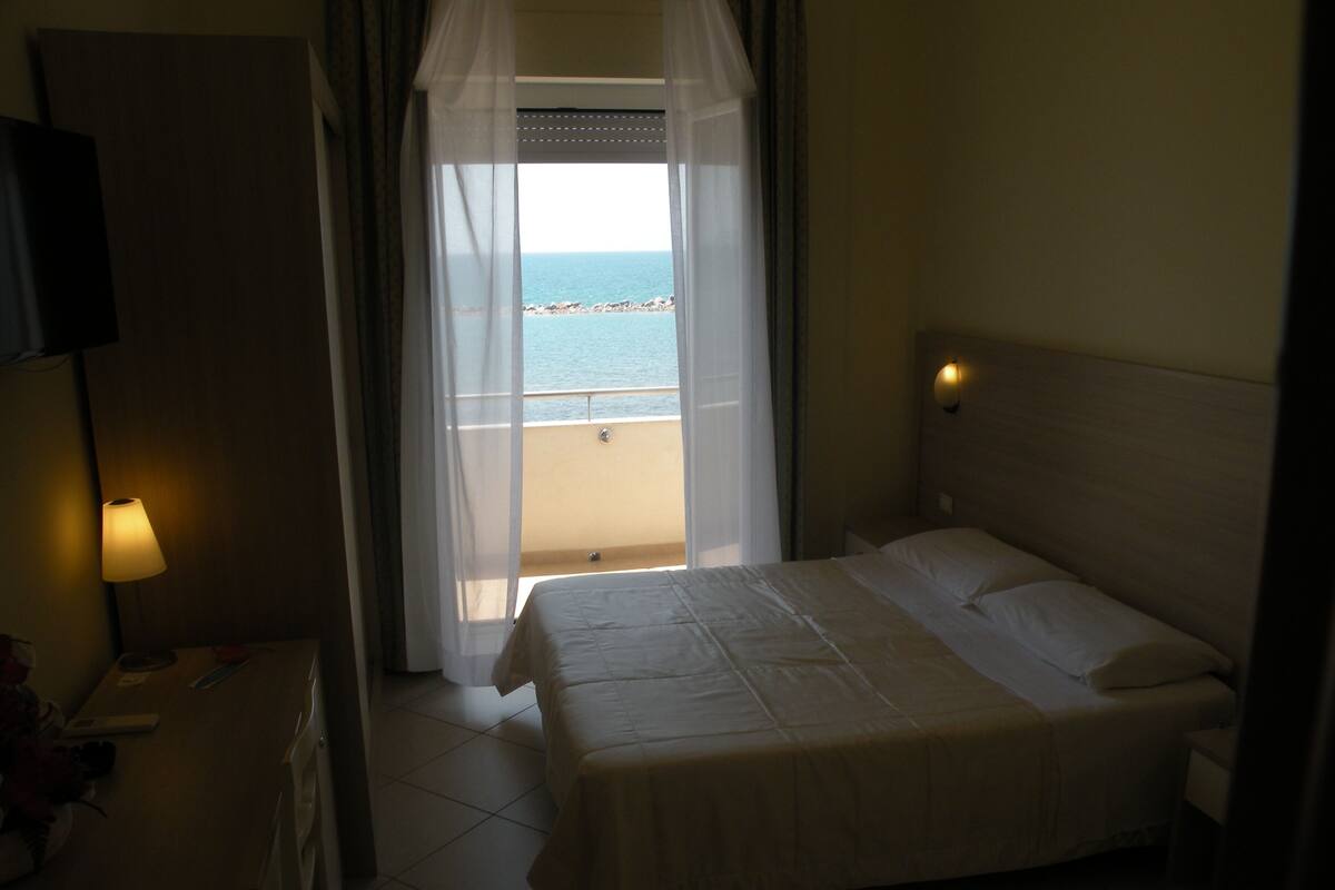 A hotel room with a bed, a bedside lamp, a view of the sea through a window, and a curtain.