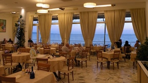 Ein Restaurant mit runden Tischen und Stühlen, großen Fenstern mit Vorhängen und Blick aufs Meer.