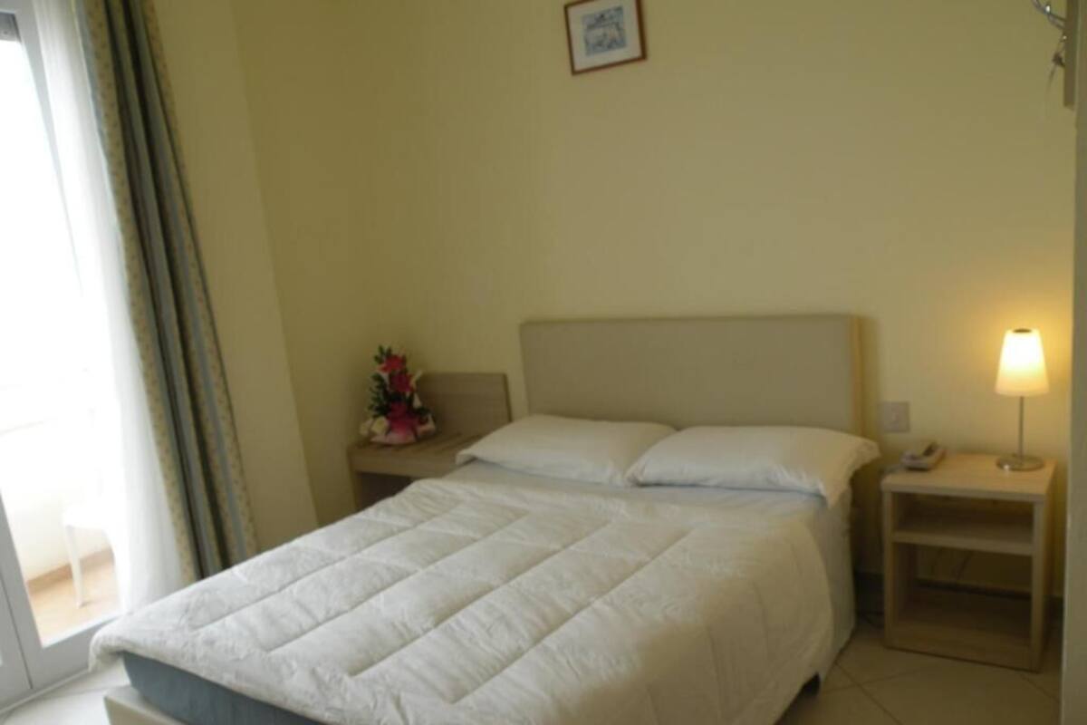 A neatly made bed with a white duvet, a bedside lamp, and a small table with a phone and a vase of flowers.