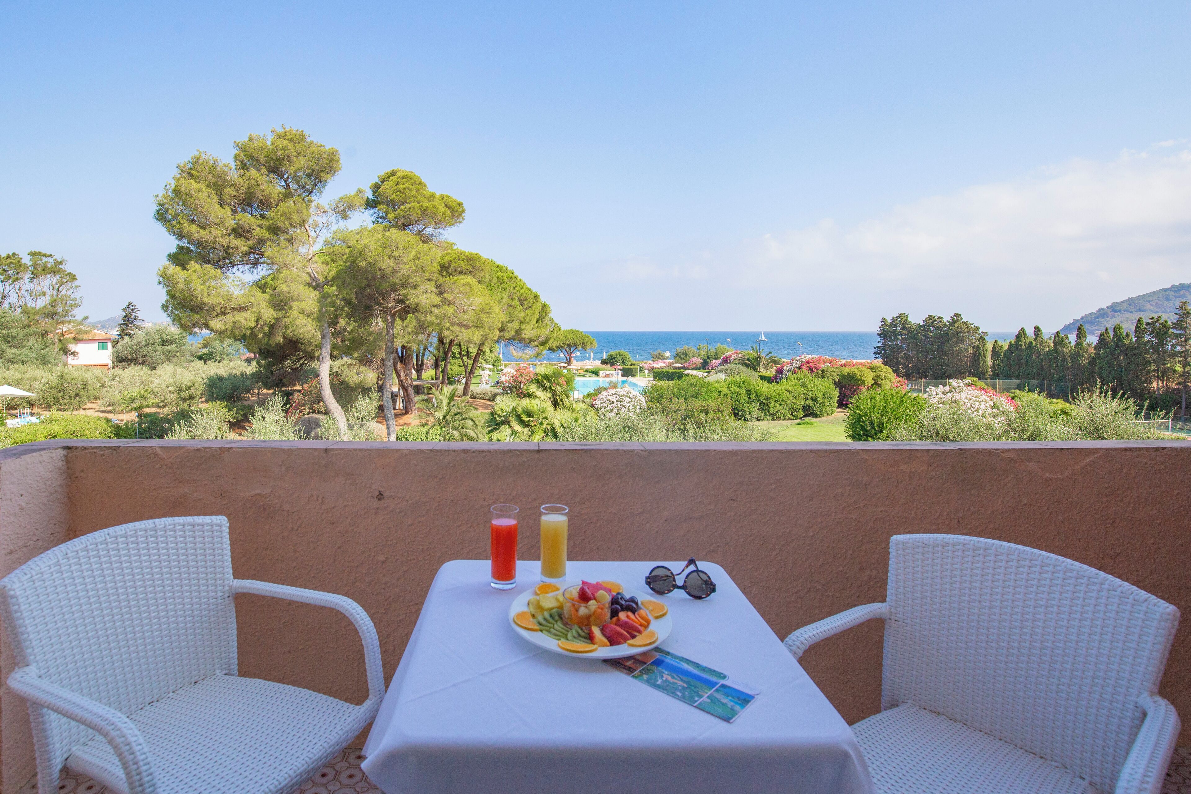 Un balcon avec deux fauteuils en osier blancs, une table sur laquelle se trouve une assiette de fruits, deux boissons et une paire de lunettes de soleil.
