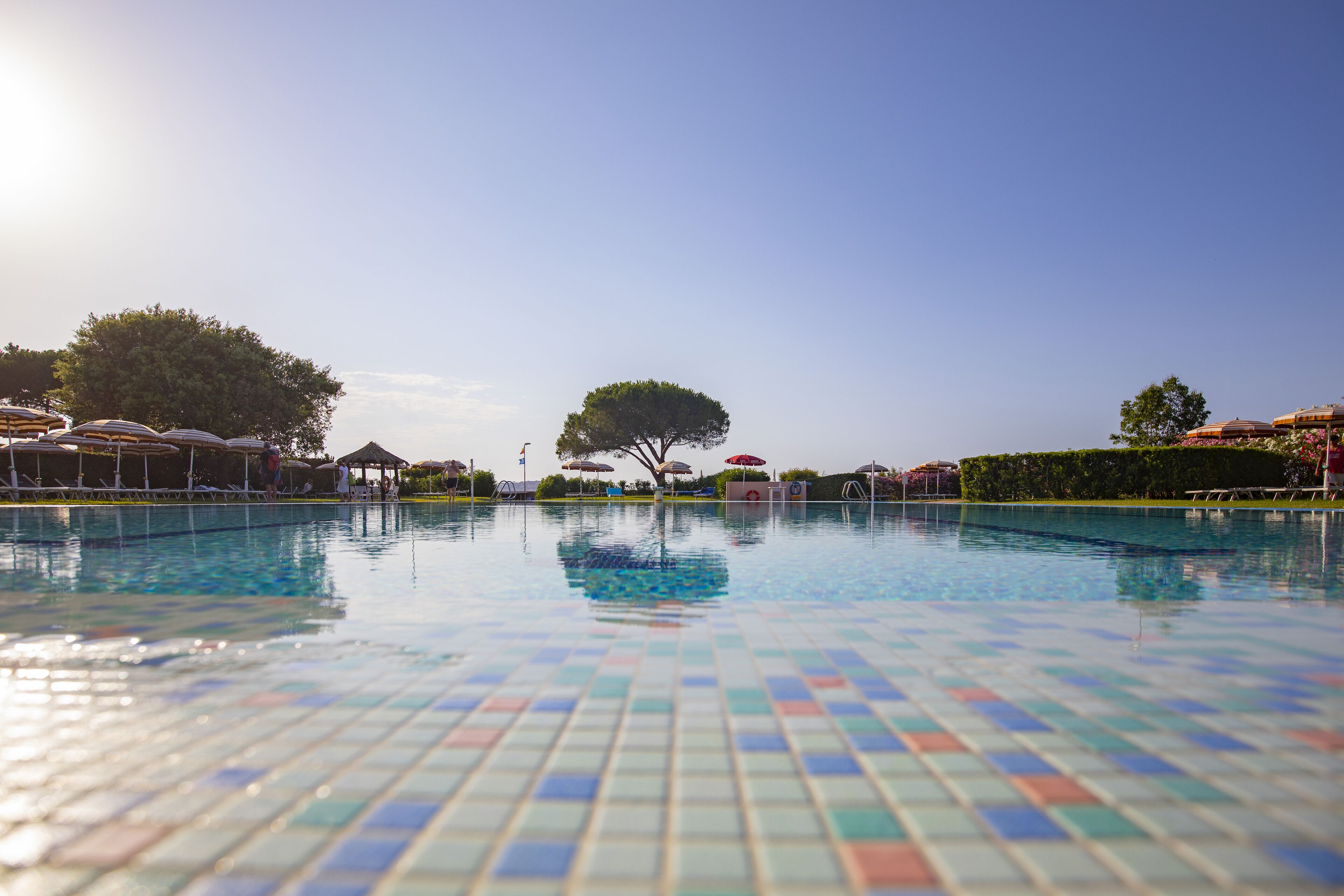 A swimming pool with tiled flooring, surrounded by umbrellas and trees.