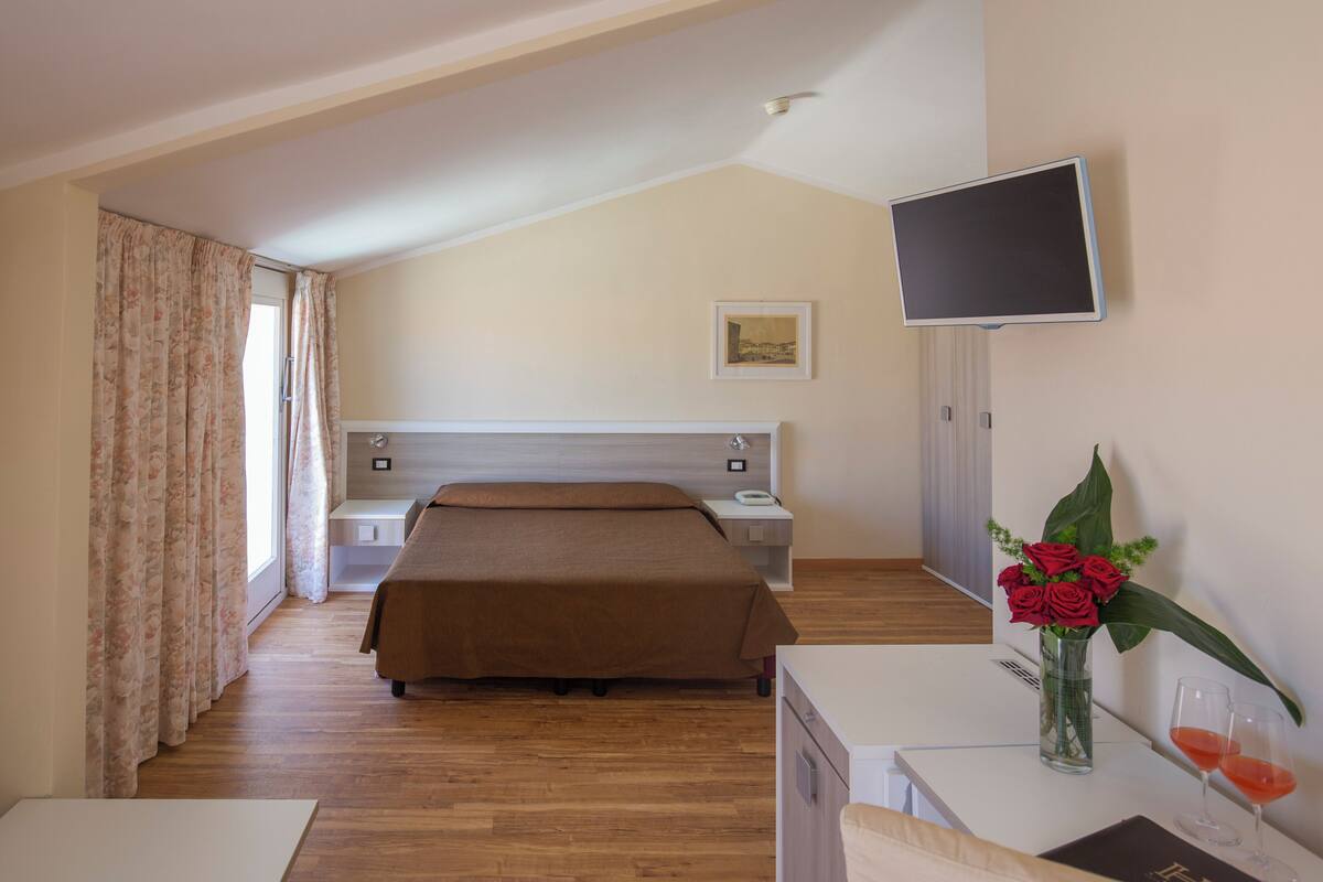 A bedroom with a bed, bedside tables, a TV, a desk with a vase of flowers and two glasses of red wine, and a window with curtains.