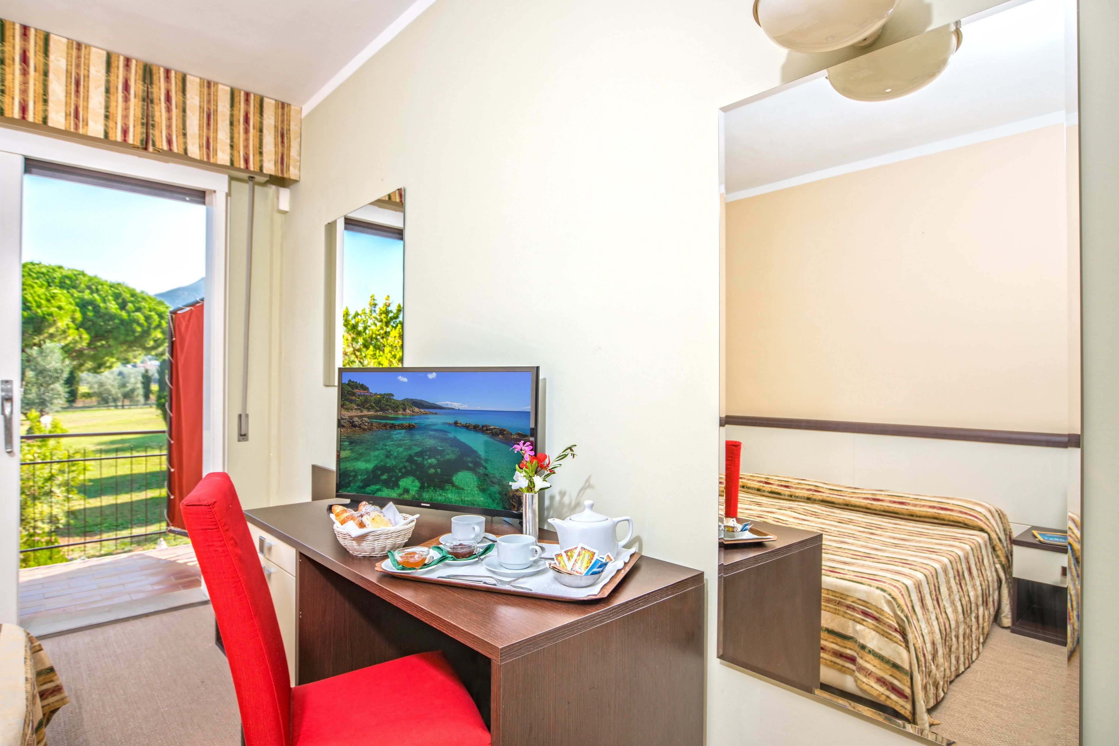 Habitación de hotel con escritorio, cama, televisor y balcón con vistas a zonas verdes y agua.