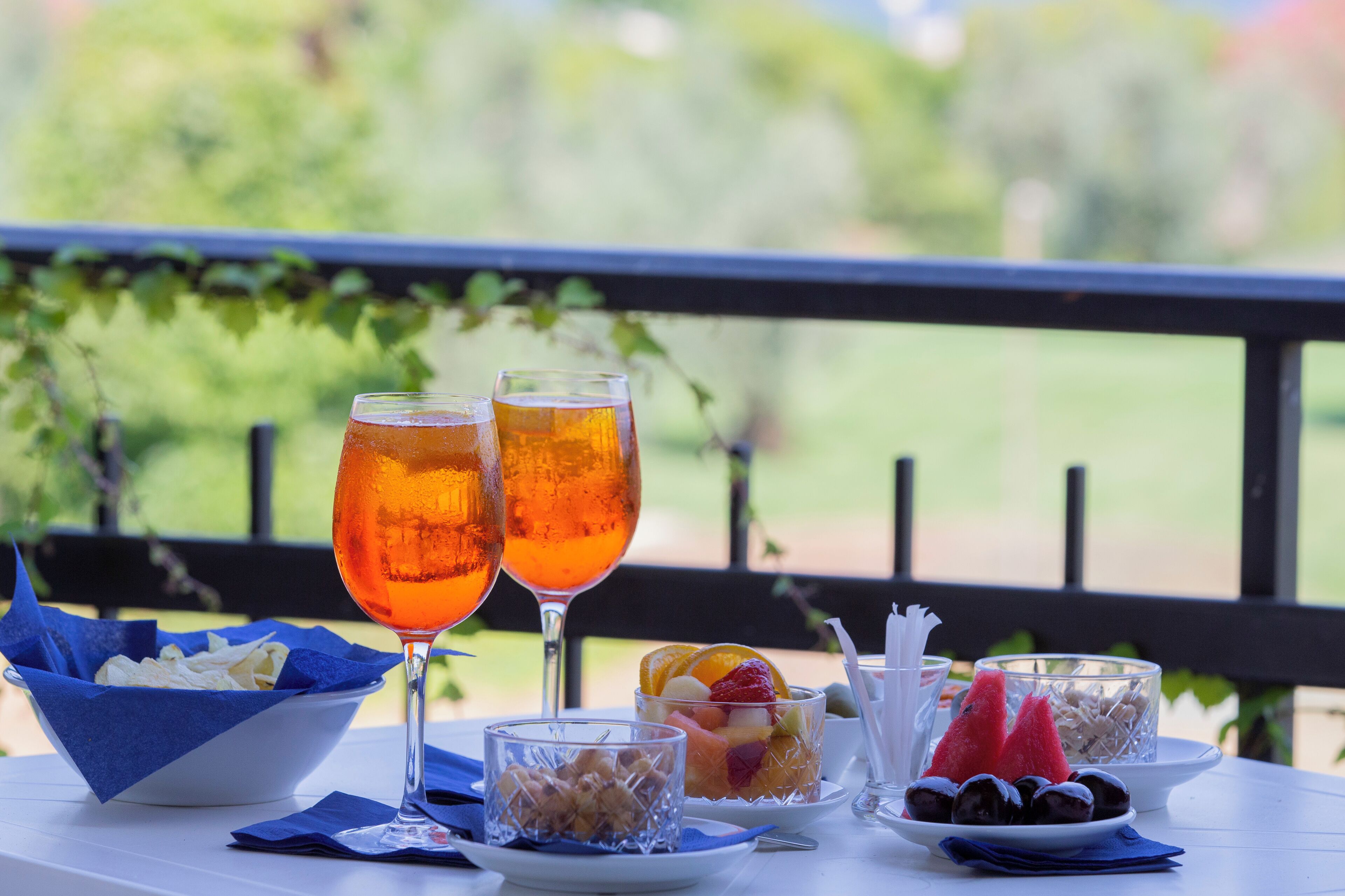 Una mesa con dos vasos de líquido ámbar, fuentes de aperitivos y un plato de fruta en un balcón con vista a follaje.