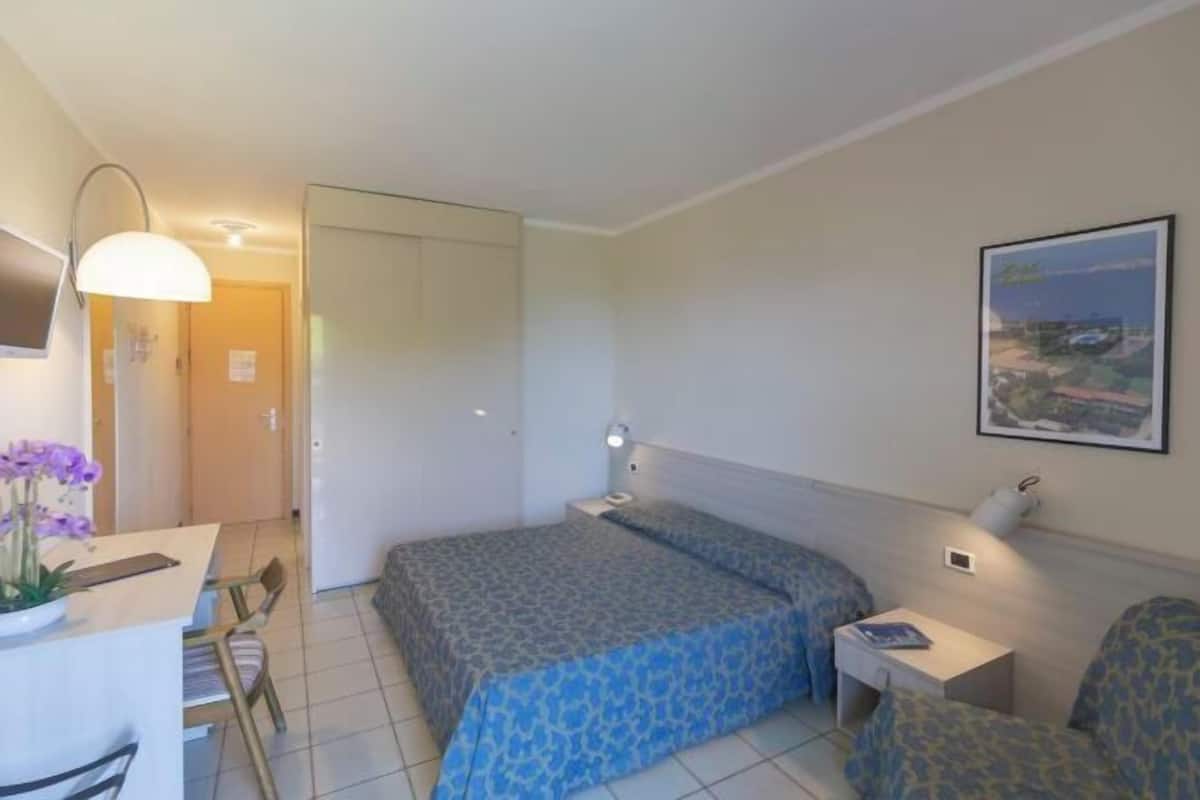 A hotel room with a bed, a desk, a chair, a lamp, and a framed picture on the wall.
