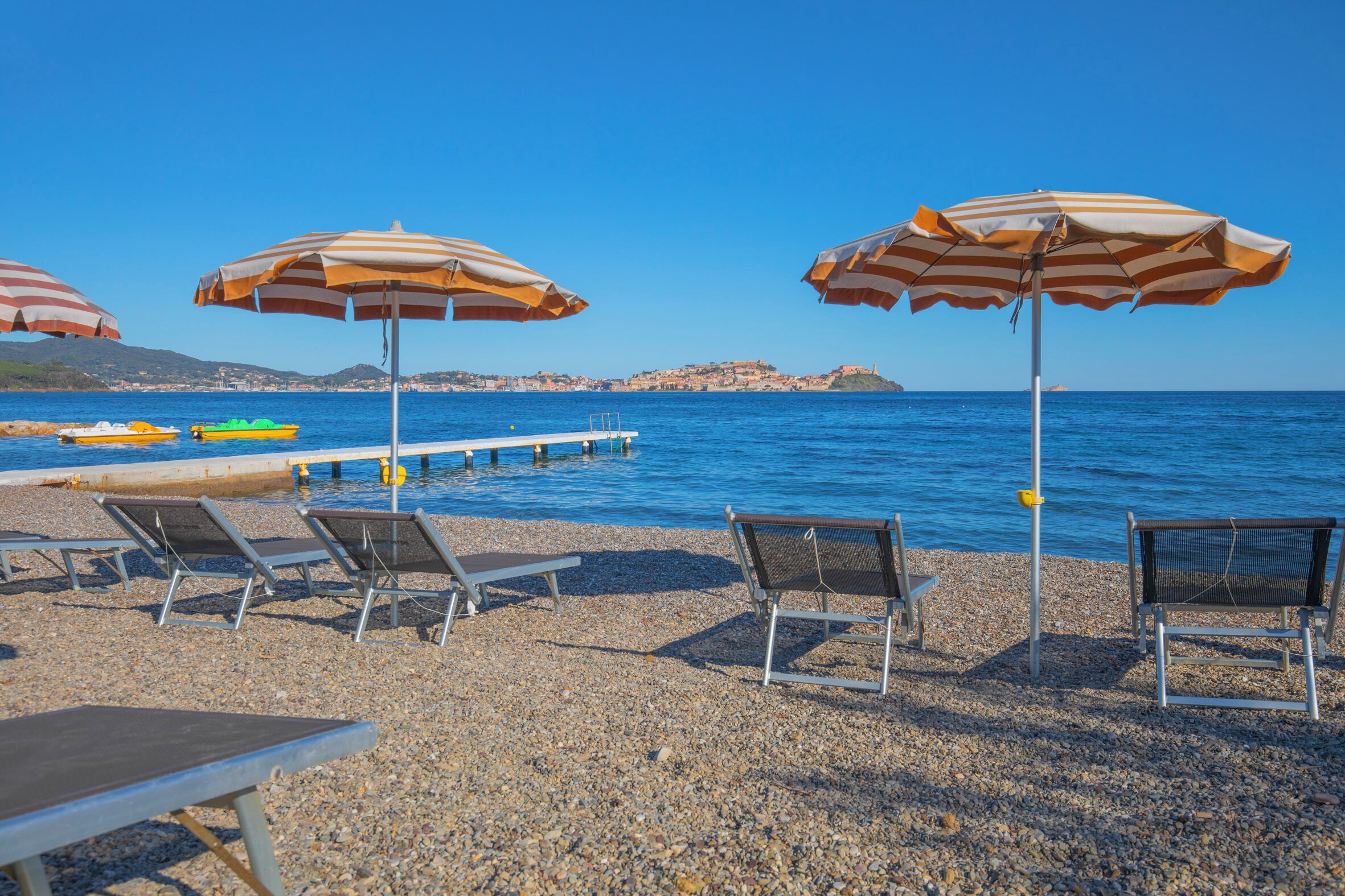 A pebble beach with lounge chairs and striped umbrellas, a dock with boats, and a clear view of the sea and distant hills.